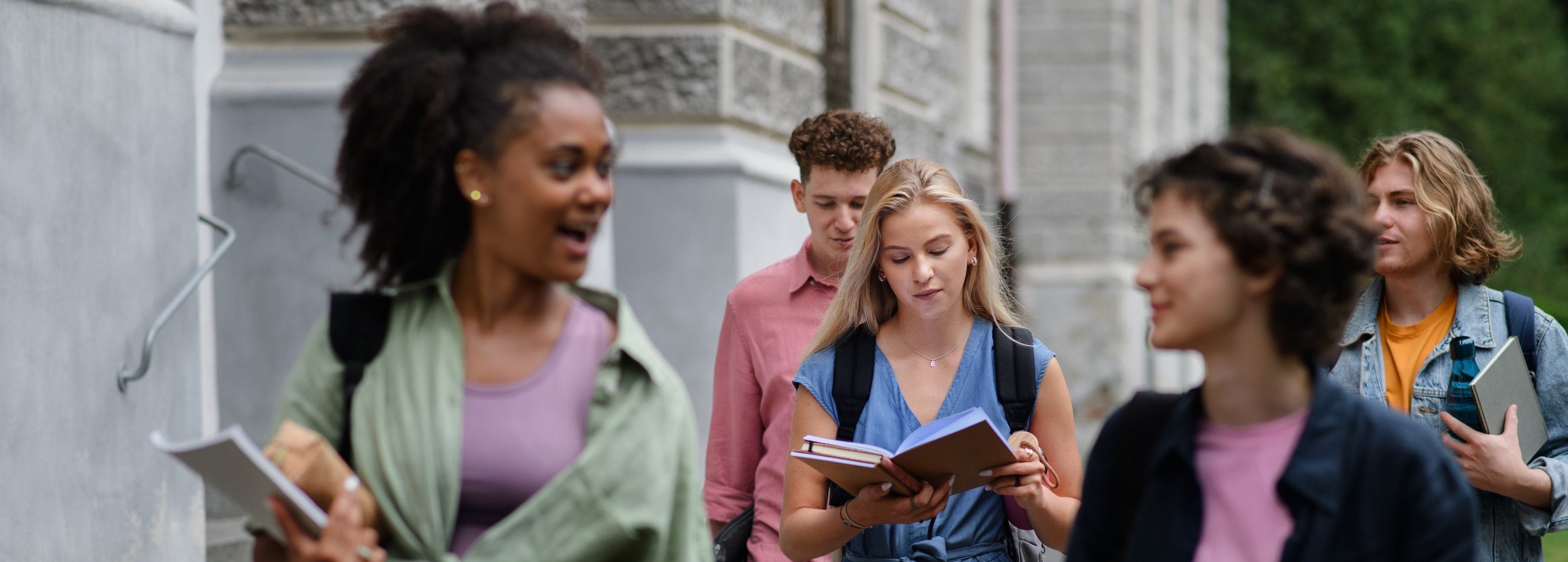 Group of happy teenagers walking together, holding books, learning, and talking in front of school.